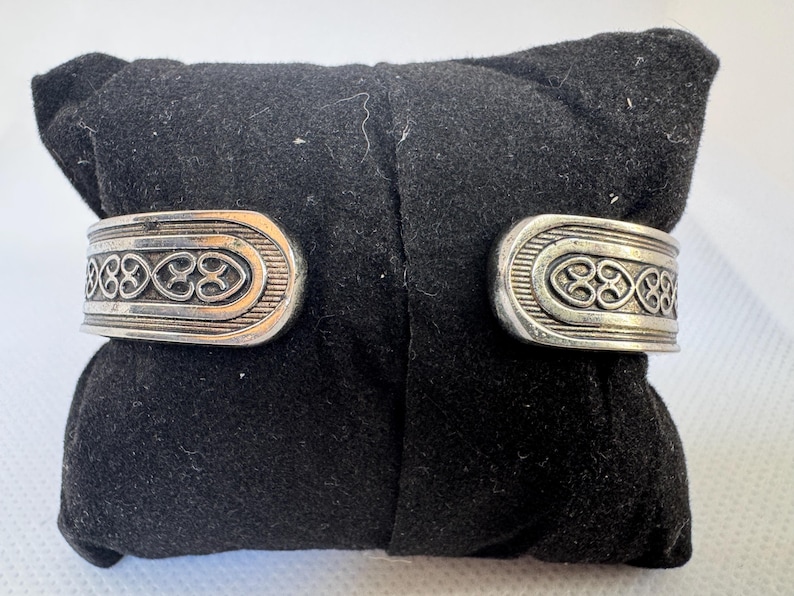 Puede incluir: Brazalete plateado con dise&ntilde;o abierto. El brazalete presenta un patr&oacute;n de nudo celta en relieve en la parte delantera. El brazalete se muestra sobre una superficie de terciopelo negro.