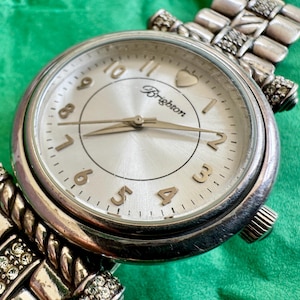 May include: A silver-tone analog wristwatch with a round face, featuring a metallic silver dial, gold-tone numerals, and the Brighton logo. The watch has a textured metal link bracelet accented with small clear crystals and a heart icon at the top.