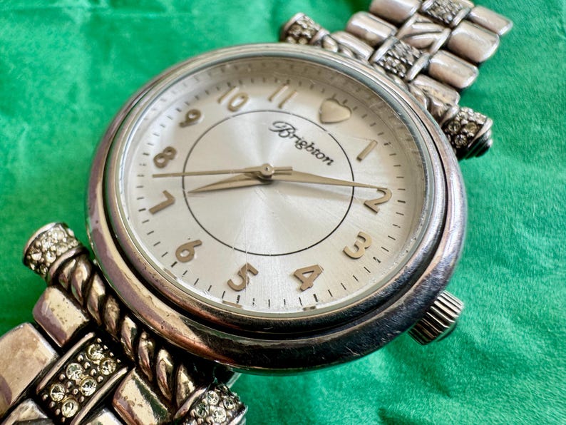 May include: A silver-tone analog wristwatch with a round face, featuring a metallic silver dial, gold-tone numerals, and the Brighton logo. The watch has a textured metal link bracelet accented with small clear crystals and a heart icon at the top.