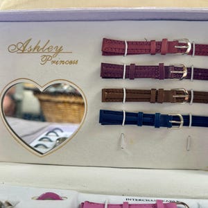 May include: A light purple presentation box labeled Ashley Princess containing four interchangeable watch straps in magenta, dark purple, brown, and blue. The box features a heart-shaped mirror on the left side and includes additional watch components.