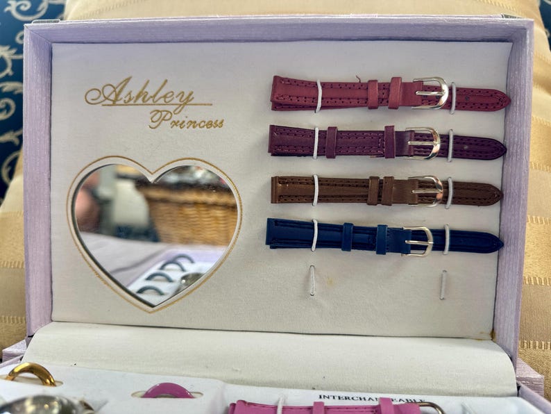May include: A light purple presentation box labeled Ashley Princess containing four interchangeable watch straps in magenta, dark purple, brown, and blue. The box features a heart-shaped mirror on the left side and includes additional watch components.