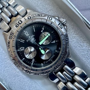 May include: A silver-tone Fossil Blue wristwatch with a black dial, featuring luminous hands, a rotating bezel with compass markings, and a multi-link metal bracelet. The watch face displays day and date sub-dials and is marked 50 meters.