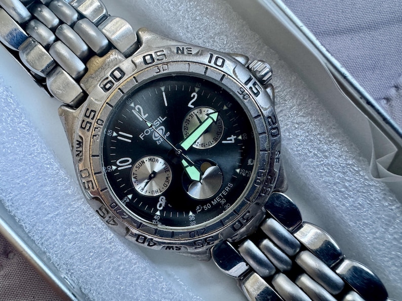 May include: A silver-tone Fossil Blue wristwatch with a black dial, featuring luminous hands, a rotating bezel with compass markings, and a multi-link metal bracelet. The watch face displays day and date sub-dials and is marked 50 meters.