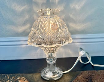 May include: A clear glass table lamp featuring a scalloped edge shade with a pressed leaf pattern and a fluted base. The lamp includes a white electrical cord with a switch and a small circular sticker on the shade and base.