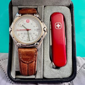May include: A silver-toned Wenger watch with a white face, black numerals, and a brown leather strap. The watch is displayed in a black case alongside a red Swiss Army knife with a white cross emblem.
