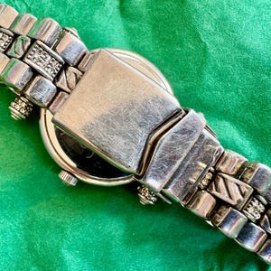 May include: A silver-tone metal wristwatch featuring a link bracelet with alternating textured diagonal lines and small clear crystal accents. The watch includes a central folding clasp mechanism and a round case with a small side crown.