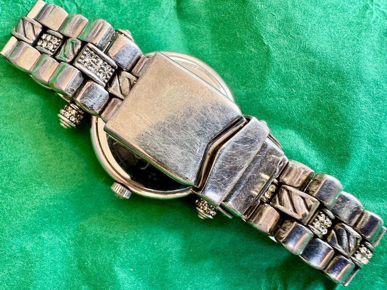 May include: A silver-tone metal wristwatch featuring a link bracelet with alternating textured diagonal lines and small clear crystal accents. The watch includes a central folding clasp mechanism and a round case with a small side crown.