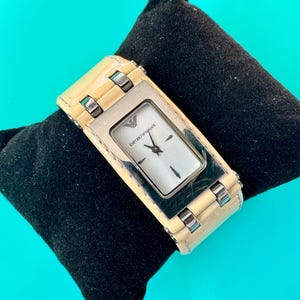 May include: Cream-colored rectangular wristwatch with a silver face and a cream leather band. The watch has a silver case and is resting on a black pillow. The watch face has the brand name "Emporio Armani".