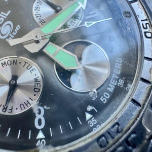 May include: A Fossil Blue analog watch with a black dial, silver-tone hands with luminous green accents, and a day-of-the-week sub-dial. The watch face features the Fossil Blue logo, a moon phase indicator, and a bezel marked with 50 meters.