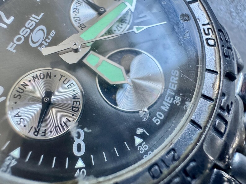 May include: A Fossil Blue analog watch with a black dial, silver-tone hands with luminous green accents, and a day-of-the-week sub-dial. The watch face features the Fossil Blue logo, a moon phase indicator, and a bezel marked with 50 meters.