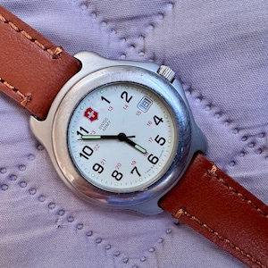 May include: A Swiss Army wristwatch with a round silver-tone metal case, a white dial featuring black Arabic numerals, red 24-hour markings, a date window at the three o'clock position, and a brown leather strap with visible stitching.