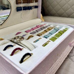 May include: A light pink rectangular storage box containing a silver wristwatch with a white strap, multiple interchangeable watch straps in various colors including pink, purple, gold, blue, and green, and several colorful circular watch bezels.