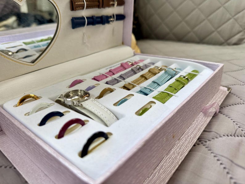 May include: A light pink rectangular storage box containing a silver wristwatch with a white strap, multiple interchangeable watch straps in various colors including pink, purple, gold, blue, and green, and several colorful circular watch bezels.