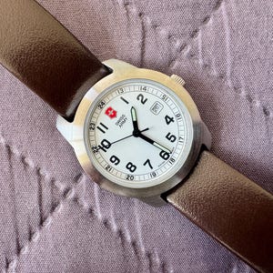 K&ouml;nnte beinhalten: Eine Swiss Army Armbanduhr mit rundem Edelstahlgeh&auml;use, wei&szlig;em Zifferblatt mit schwarzen arabischen Ziffern und braunem Lederarmband. Das Zifferblatt zeigt das Swiss Army Logo, ein Datumsfenster auf der Drei-Uhr-Position und eine 24-Stunden-Skala.