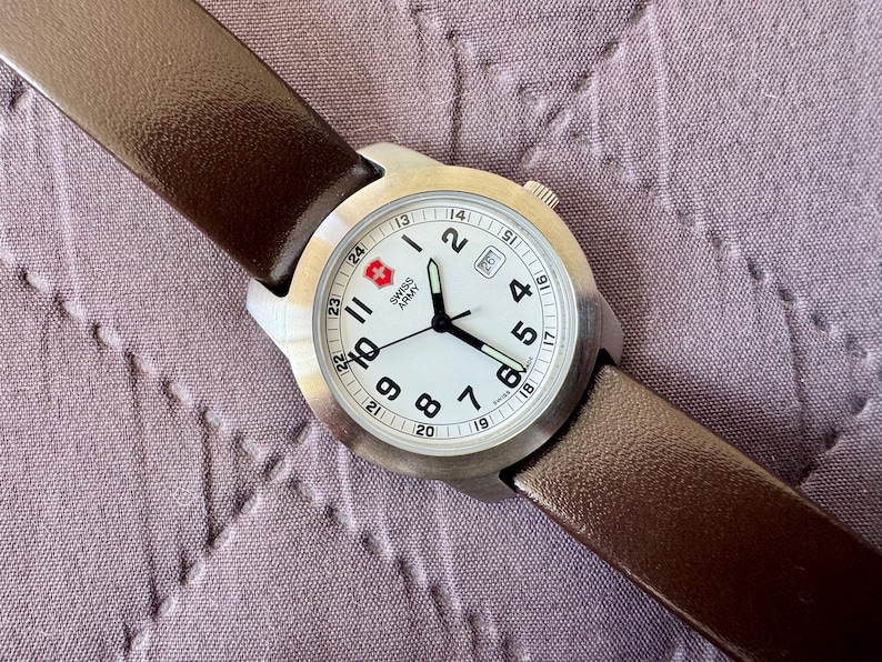 K&ouml;nnte beinhalten: Eine Swiss Army Armbanduhr mit rundem Edelstahlgeh&auml;use, wei&szlig;em Zifferblatt mit schwarzen arabischen Ziffern und braunem Lederarmband. Das Zifferblatt zeigt das Swiss Army Logo, ein Datumsfenster auf der Drei-Uhr-Position und eine 24-Stunden-Skala.