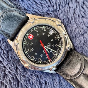 K&ouml;nnte beinhalten: Eine silberfarbene Armbanduhr mit schwarzem Zifferblatt, wei&szlig;en Ziffern und einem roten Sekundenzeiger. Die Uhr hat eine Datumsanzeige bei 3 Uhr und ein schwarzes Lederarmband. Das Zifferblatt tr&auml;gt den Schriftzug "WENGER" und ein rotes Kreuz-Logo.