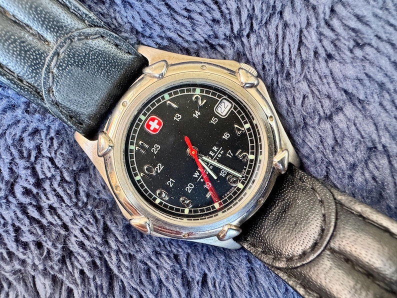 K&ouml;nnte beinhalten: Eine silberfarbene Armbanduhr mit schwarzem Zifferblatt, wei&szlig;en Ziffern und einem roten Sekundenzeiger. Die Uhr hat eine Datumsanzeige bei 3 Uhr und ein schwarzes Lederarmband. Das Zifferblatt tr&auml;gt den Schriftzug "WENGER" und ein rotes Kreuz-Logo.