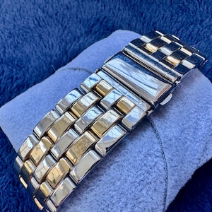May include: A two-tone metal watch band with silver and gold links. The watch band is resting on a small, light blue pillow. The background is a dark blue, textured fabric.