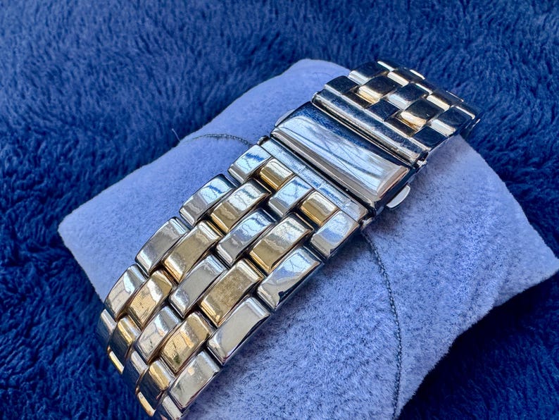May include: A two-tone metal watch band with silver and gold links. The watch band is resting on a small, light blue pillow. The background is a dark blue, textured fabric.