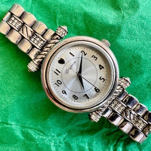 May include: A silver-tone analog wristwatch featuring a round face with black numerals, a heart icon at the twelve o'clock position, and the Brighton logo. The metal link band is accented with small clear crystals and a twisted rope-style design.