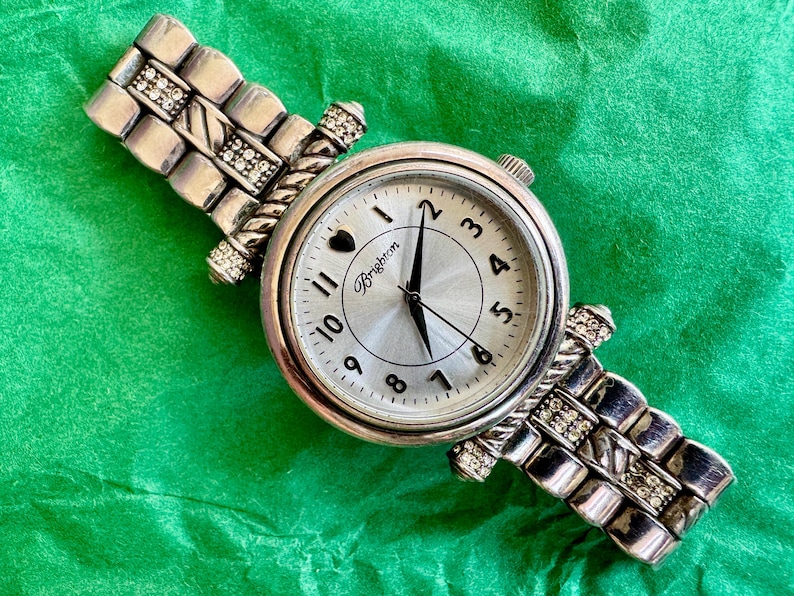 May include: A silver-tone analog wristwatch featuring a round face with black numerals, a heart icon at the twelve o'clock position, and the Brighton logo. The metal link band is accented with small clear crystals and a twisted rope-style design.
