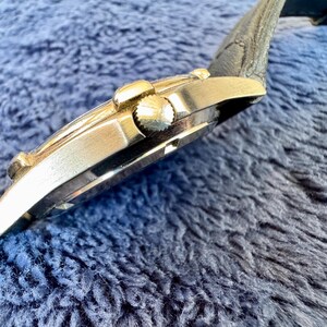K&ouml;nnte beinhalten: Seitenansicht einer goldfarbenen Armbanduhr mit schwarzem Lederarmband. Das Zifferblatt ist nicht sichtbar. Die Uhr hat eine strukturierte Krone und ein geschwungenes Design. Die Uhr befindet sich vor einem blauen, strukturierten Hintergrund.