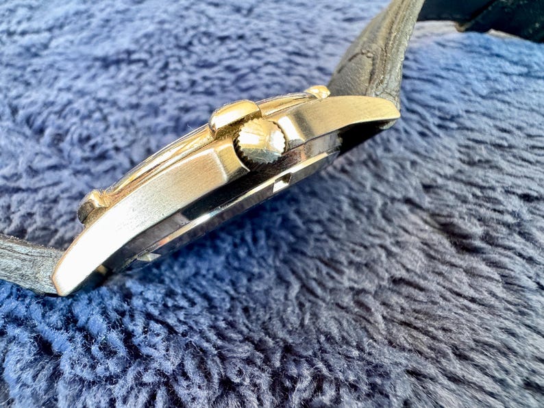 K&ouml;nnte beinhalten: Seitenansicht einer goldfarbenen Armbanduhr mit schwarzem Lederarmband. Das Zifferblatt ist nicht sichtbar. Die Uhr hat eine strukturierte Krone und ein geschwungenes Design. Die Uhr befindet sich vor einem blauen, strukturierten Hintergrund.