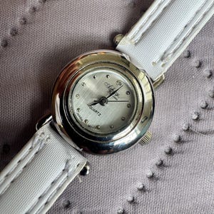 May include: A round silver-tone wristwatch with a white dial featuring the text Ashley and Quartz Japan Movt. The watch has a polished metallic bezel and a white strap with visible stitching along the edges. The dial includes small dot markers.