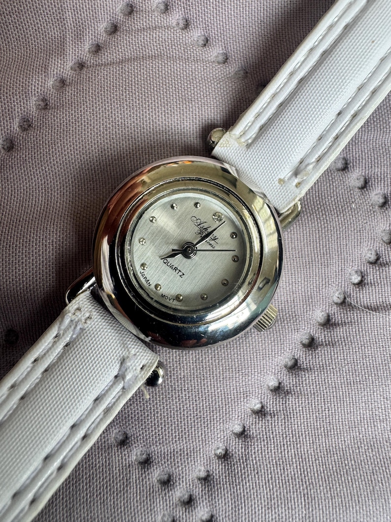 May include: A round silver-tone wristwatch with a white dial featuring the text Ashley and Quartz Japan Movt. The watch has a polished metallic bezel and a white strap with visible stitching along the edges. The dial includes small dot markers.
