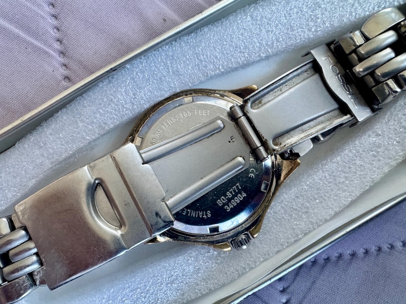 May include: A silver-tone Fossil wristwatch with a stainless steel link bracelet. The back of the watch case displays the text BQ-8777, 349904, and 50 meters/165 feet. The watch is shown from the back, revealing the metal clasp and casing.