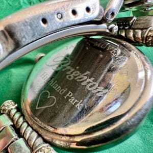 May include: A silver-tone metal wristwatch with a round case and a link bracelet. The back of the watch case is engraved with the text Brighton, Highland Park, and a heart symbol, along with the words Japan Movement.