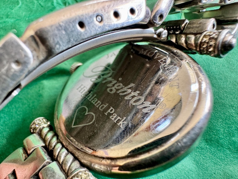 May include: A silver-tone metal wristwatch with a round case and a link bracelet. The back of the watch case is engraved with the text Brighton, Highland Park, and a heart symbol, along with the words Japan Movement.