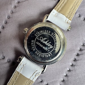 May include: A wristwatch with a round silver-tone case and a white strap. The back of the watch case is made of stainless steel and features the text Ashley, Japan Movt, and Water Resistant. The strap has visible stitching along the edges.
