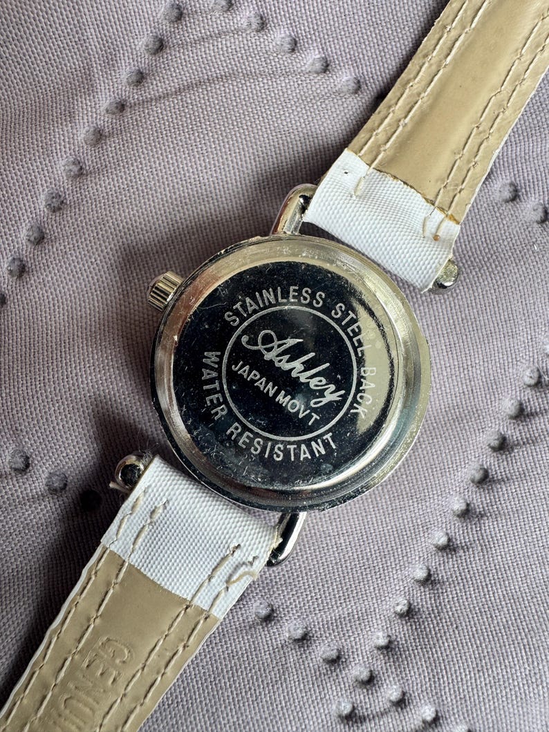 May include: A wristwatch with a round silver-tone case and a white strap. The back of the watch case is made of stainless steel and features the text Ashley, Japan Movt, and Water Resistant. The strap has visible stitching along the edges.