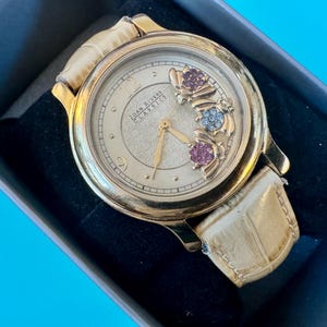 May include: Gold wristwatch with a cream face and a light leather strap. The watch face has floral detailing with pink, blue, and purple gemstones. The watch is presented in a black box.