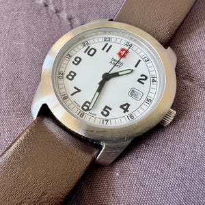 K&ouml;nnte beinhalten: Eine Swiss Army Armbanduhr mit rundem Edelstahlgeh&auml;use, wei&szlig;em Zifferblatt mit schwarzen arabischen Ziffern, einer 24-Stunden-Skala, Datumsanzeige auf der Drei-Uhr-Position, leuchtenden Zeigern und einem glatten, braunen Lederarmband.