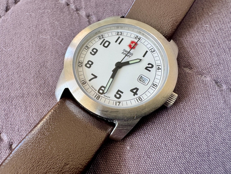 K&ouml;nnte beinhalten: Eine Swiss Army Armbanduhr mit rundem Edelstahlgeh&auml;use, wei&szlig;em Zifferblatt mit schwarzen arabischen Ziffern, einer 24-Stunden-Skala, Datumsanzeige auf der Drei-Uhr-Position, leuchtenden Zeigern und einem glatten, braunen Lederarmband.