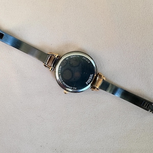 May include: A Fossil brand wristwatch with a black stainless steel band and gold-tone accents. The watch face is black with gold-tone hands and markers. The back of the watch is visible, with the brand name and model information.