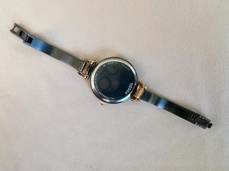 May include: A Fossil brand wristwatch with a black stainless steel band and gold-tone accents. The watch face is black with gold-tone hands and markers. The back of the watch is visible, with the brand name and model information.