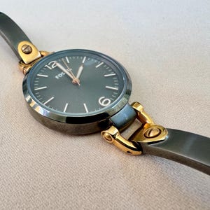 May include: A Fossil watch with a dark gray face and gold-tone accents. The watch has a dark gray band and a round face with the brand name "Fossil" printed on it. The watch is a stylish accessory.