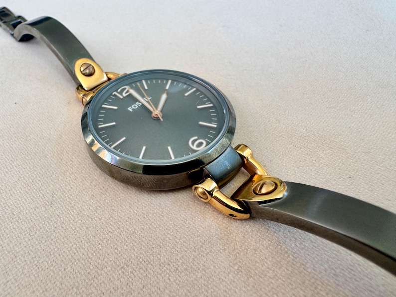 May include: A Fossil watch with a dark gray face and gold-tone accents. The watch has a dark gray band and a round face with the brand name "Fossil" printed on it. The watch is a stylish accessory.