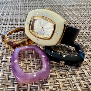 May include: A quartz wristwatch with a cream-colored rectangular case featuring four small embedded crystals, a silver dial, and a black strap. The set includes three additional interchangeable rectangular bezels in tortoise, black, and purple.