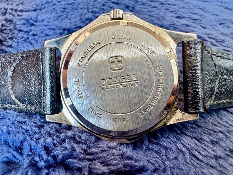K&ouml;nnte beinhalten: Nahaufnahme der R&uuml;ckseite einer Wenger SAK Design Uhr. Die Uhr hat einen Edelstahlr&uuml;cken mit dem Markennamen und Logo. Die Uhr ist bis 100 m wasserdicht und hat ein schwarzes Lederarmband.