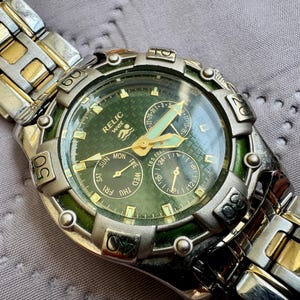 May include: A Relic Wet wristwatch featuring a green textured dial with gold-tone hands and markers. The watch has a silver and gold-tone metal link band and a rotating bezel with engraved numbers. It includes three sub-dials for date and time.