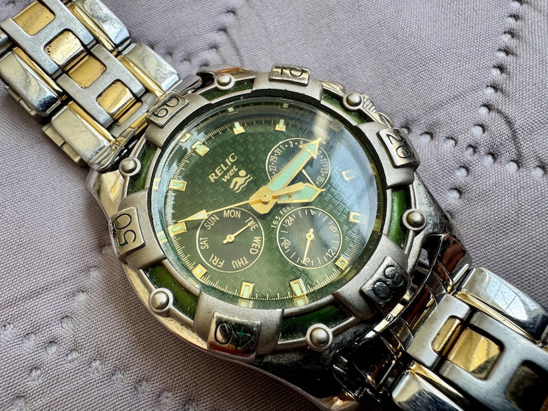 May include: A Relic Wet wristwatch featuring a green textured dial with gold-tone hands and markers. The watch has a silver and gold-tone metal link band and a rotating bezel with engraved numbers. It includes three sub-dials for date and time.