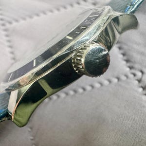 May include: A silver-toned wristwatch featuring a dark blue bezel and a matching dark blue textured leather strap. The side of the watch case displays a ridged, circular crown used for time adjustment, positioned against the polished metal housing.