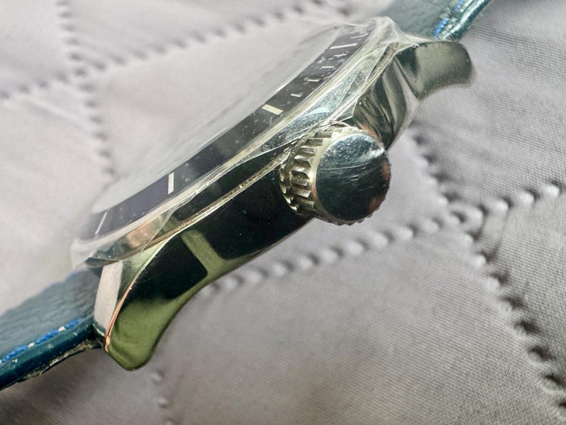 May include: A silver-toned wristwatch featuring a dark blue bezel and a matching dark blue textured leather strap. The side of the watch case displays a ridged, circular crown used for time adjustment, positioned against the polished metal housing.