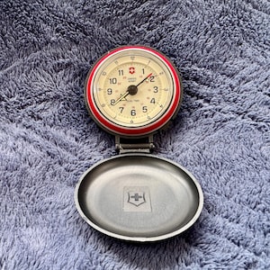 May include: A Swiss Army pocket watch with a red bezel and a cream-colored face. The watch has black numerals and hands, and the case is open, revealing a silver-colored interior with a Swiss Army emblem.