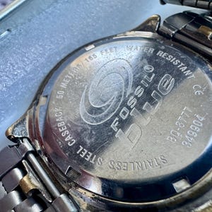 May include: The stainless steel caseback of a Fossil Blue watch featuring an engraved logo, the text 50 METERS/165 FEET WATER RESISTANT, the model number BQ-8777, and serial number 349904, attached to a metallic link watch band with gold-tone accents.