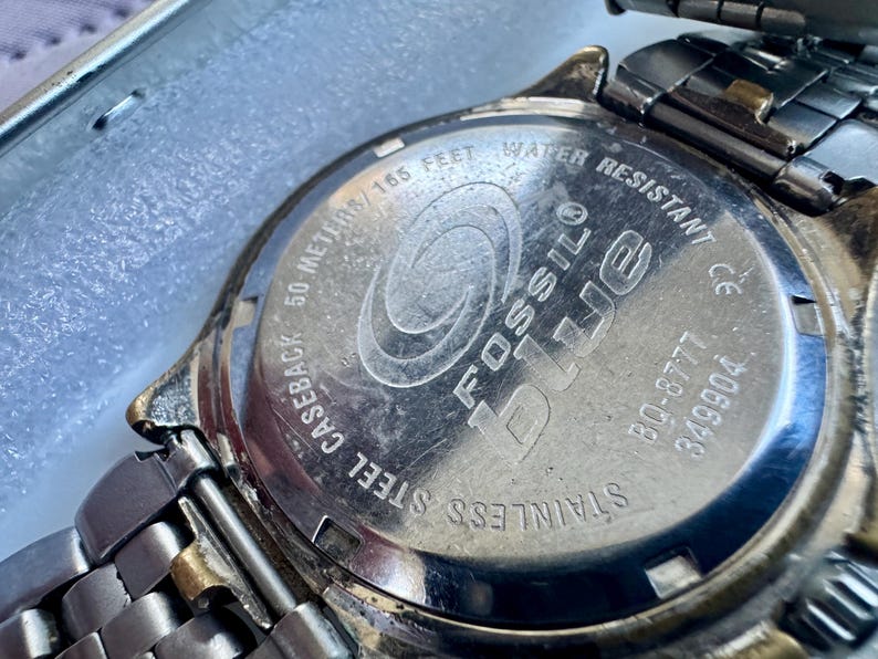 May include: The stainless steel caseback of a Fossil Blue watch featuring an engraved logo, the text 50 METERS/165 FEET WATER RESISTANT, the model number BQ-8777, and serial number 349904, attached to a metallic link watch band with gold-tone accents.